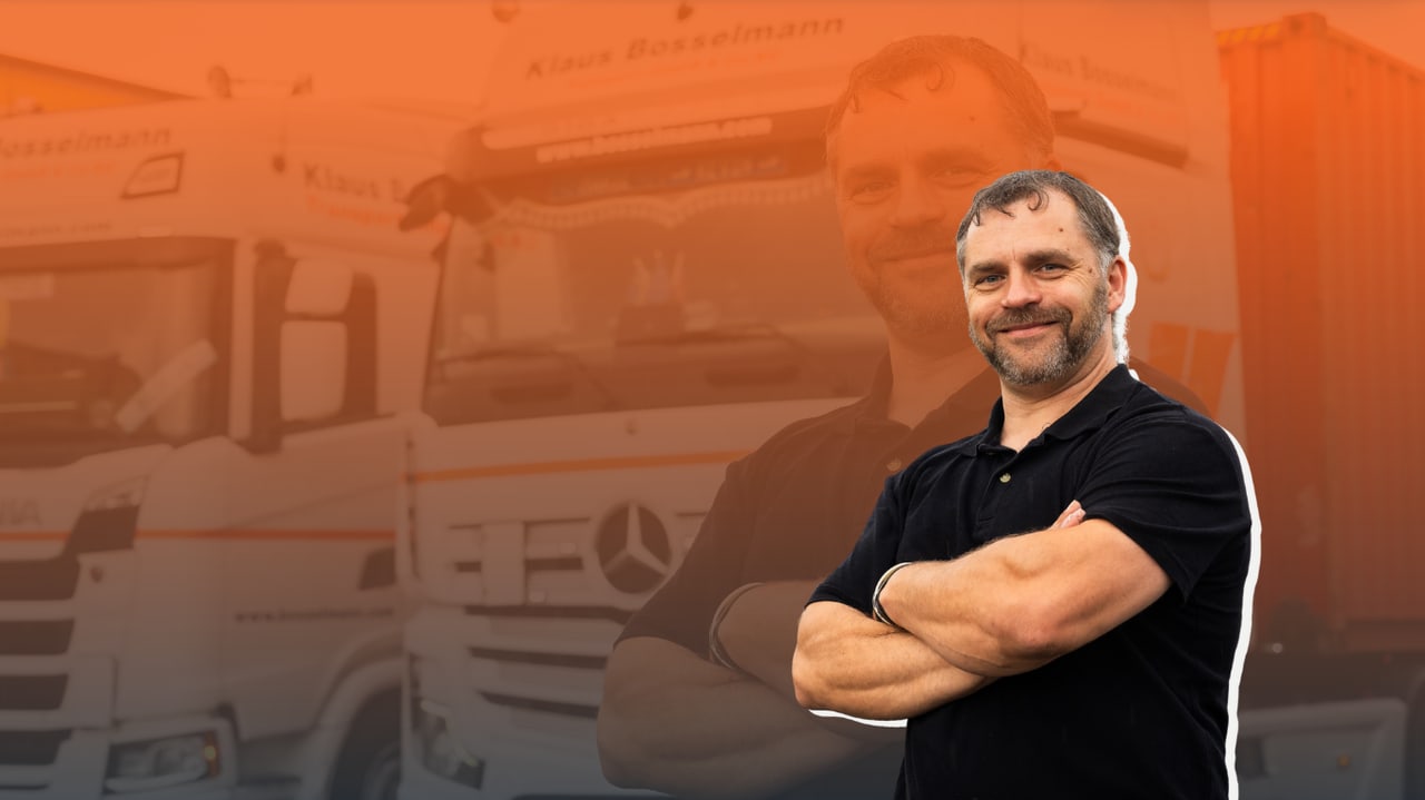 Ein lächelnder Mann mit verschränkten Armen vor einem unscharfen Bild von zwei Lastwagen.