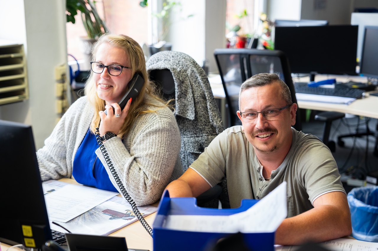 Zwei Büroangestellte an Schreibtischen; eine Frau telefoniert, ein Mann lächelt in die Kamera.