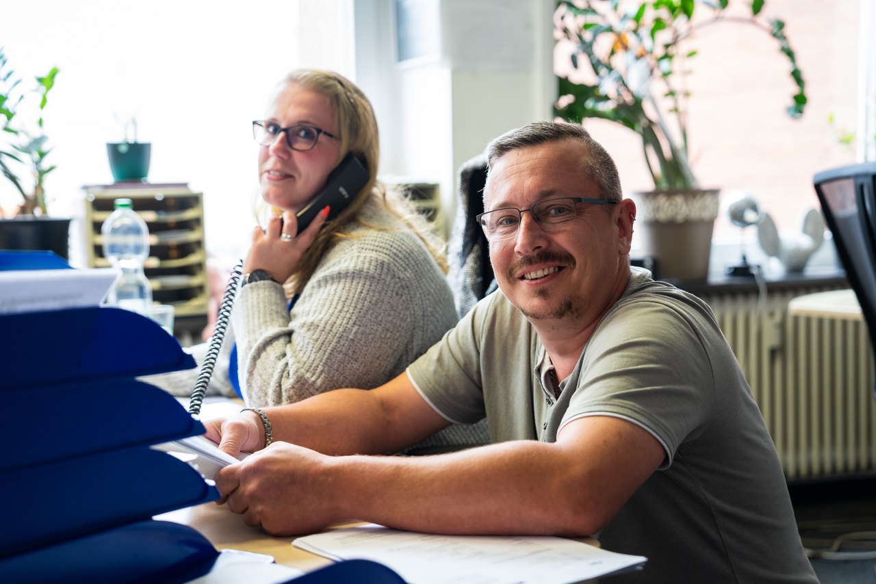 Zwei lächelnde Mitarbeiter sitzen an Schreibtischen im Büro, eine telefoniert, der andere blickt in die Kamera.