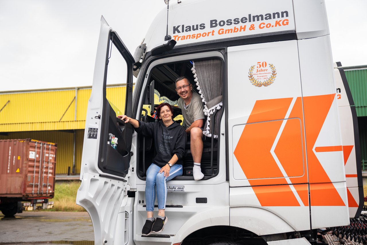 Zwei Männer posieren bei einem weißen LKW; einer sitzt im Fahrerhaus, der andere steht davor.